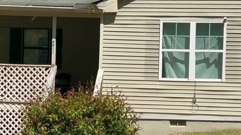 El frente de una casa de tablillas claras muestra una ventana con cortinas y un porche con rejas de madera.