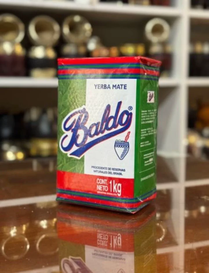 Un paquete de yerba mate Baldo de un kilo apoyado sobre una superficie brillante con mates al fondo.