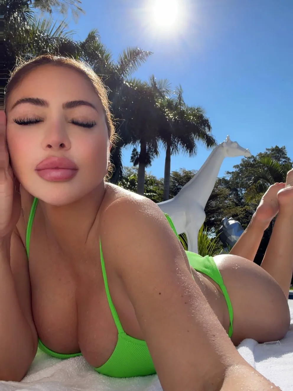 Larsa Pippen toma sol recostada boca abajo en bikini verde flúo cerca de una escultura de jirafa blanca.