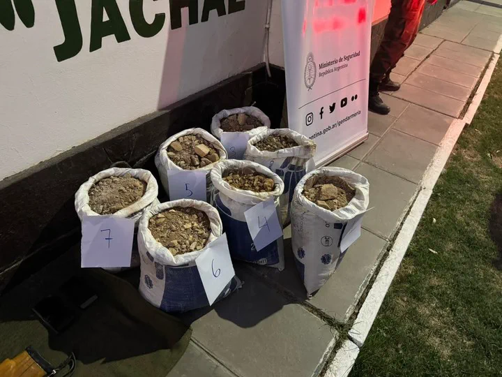 Bolsas blancas numeradas con piedras y minerales en la vereda junto a un cartel institucional.