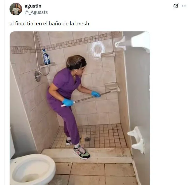 Una mujer con ambo violeta y guantes limpia el piso y los azulejos de una ducha con un cepillo de mango largo.