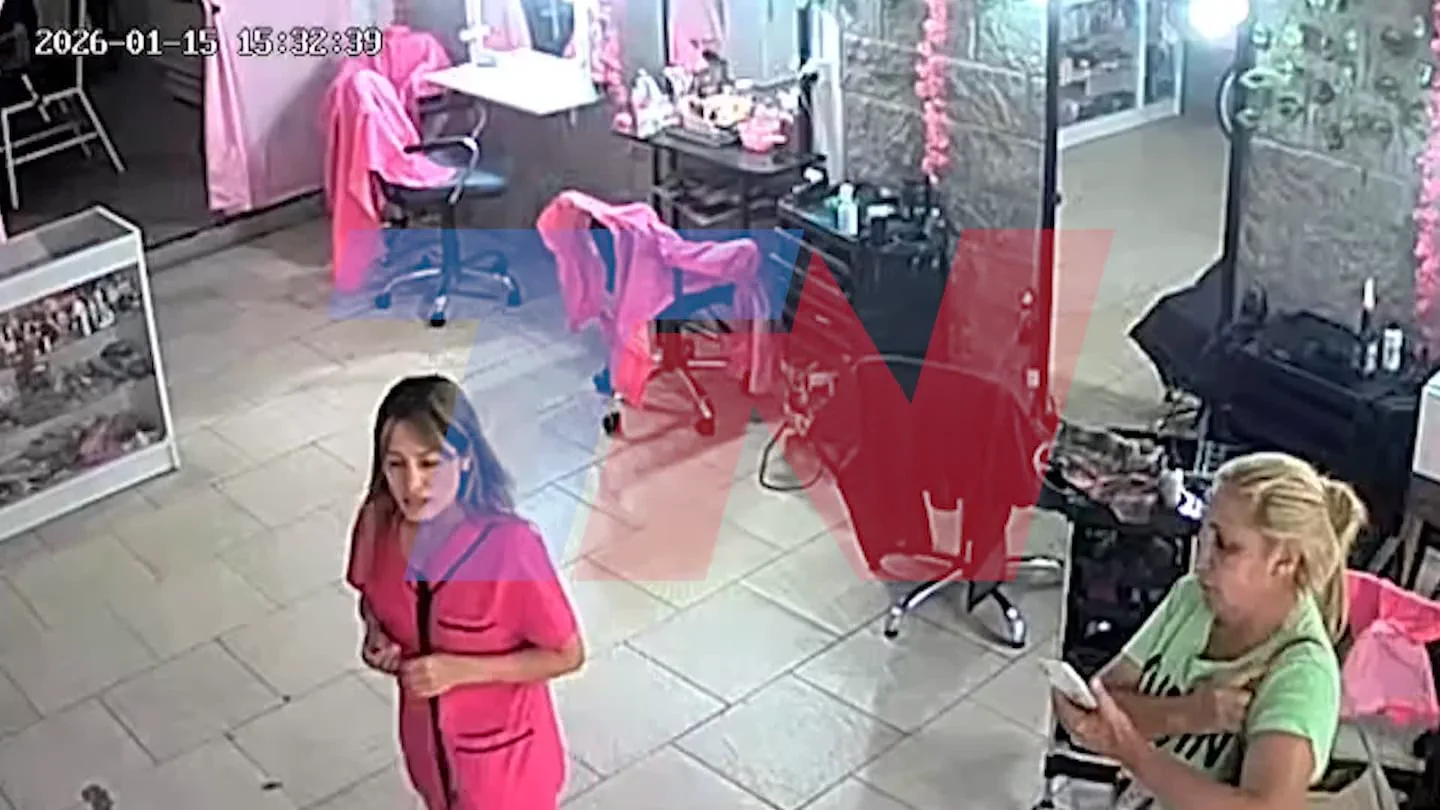 Captura de cámara de seguridad dentro de una peluquería con dos mujeres de pie, una en ambo fucsia y otra con remera verde.