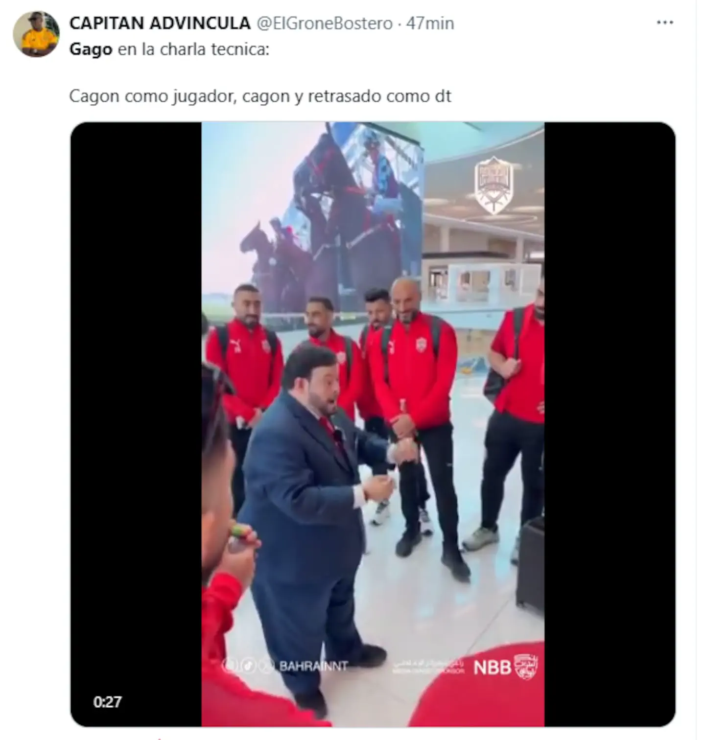 Un hombre de traje habla gesticulando ante varios hombres vestidos con camperas deportivas rojas en un hall.