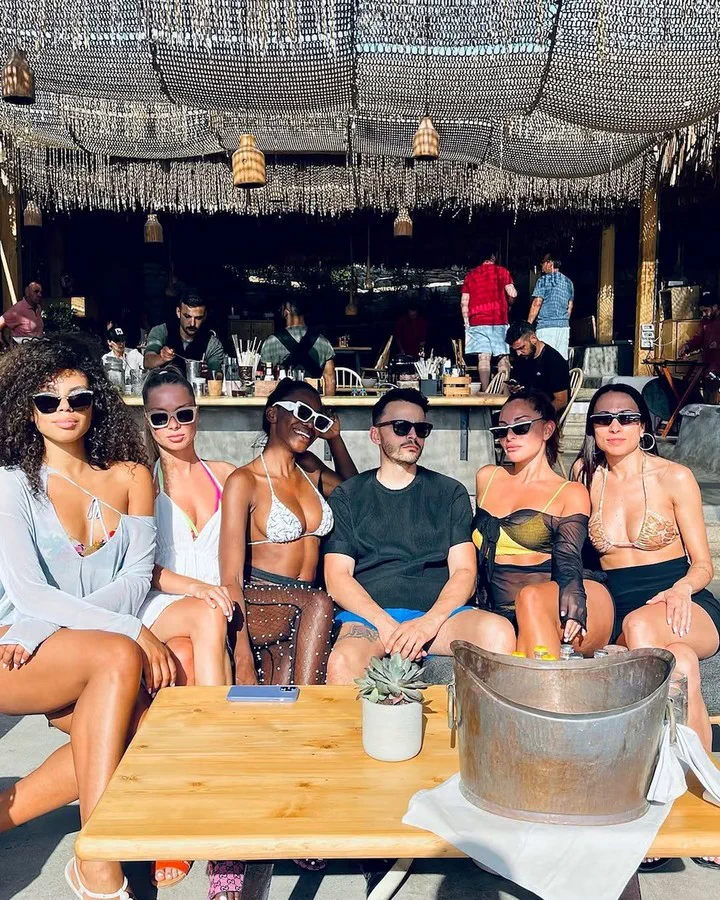 Varias personas con anteojos de sol posan sentadas alrededor de una mesa baja en un parador al aire libre.