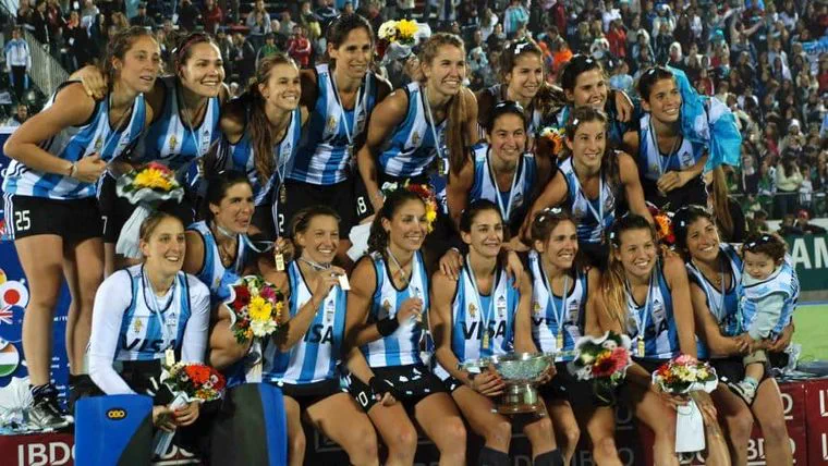 Jugadoras de la selección argentina de hockey posan sonrientes con medallas, flores y un trofeo en una cancha.