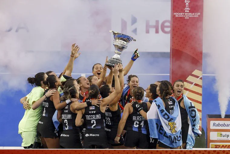 Un grupo de jugadoras de hockey con uniforme negro y celeste festejan levantando un trofeo.