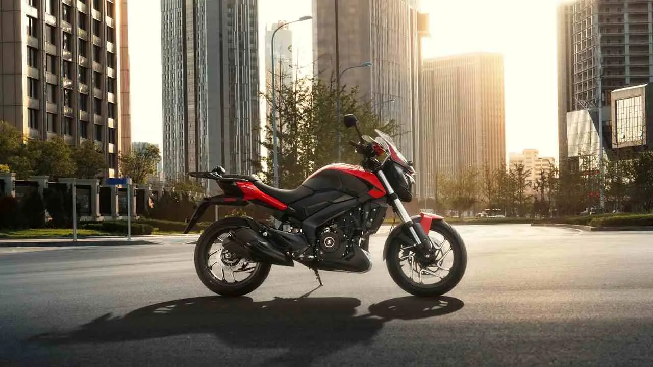 Una moto roja y negra se encuentra parada sobre el asfalto de una avenida con edificios al atardecer.