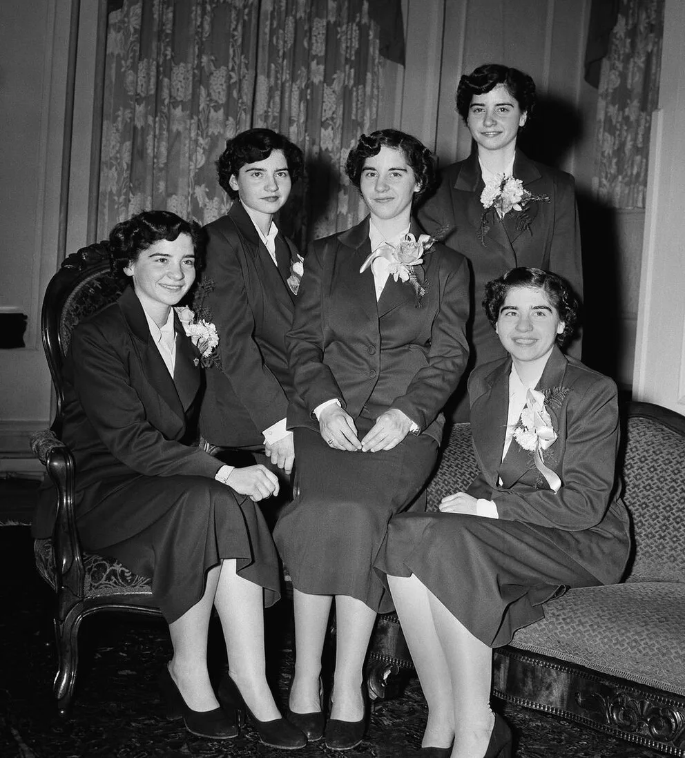Las quintillizas Dionne posan sonrientes con trajes oscuros y flores en una foto en blanco y negro.