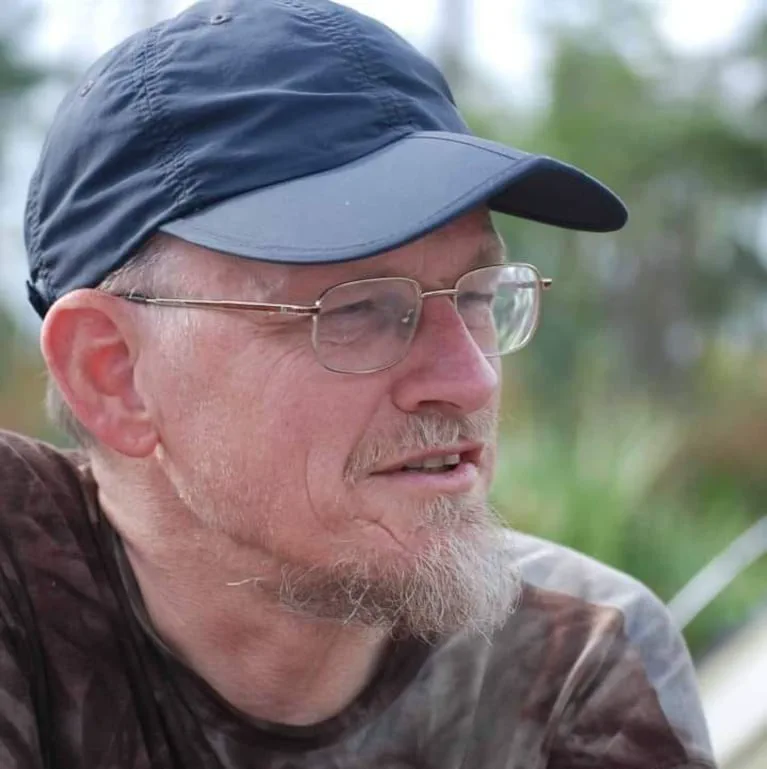 Un hombre de perfil con anteojos, barba perilla y gorra azul al aire libre.