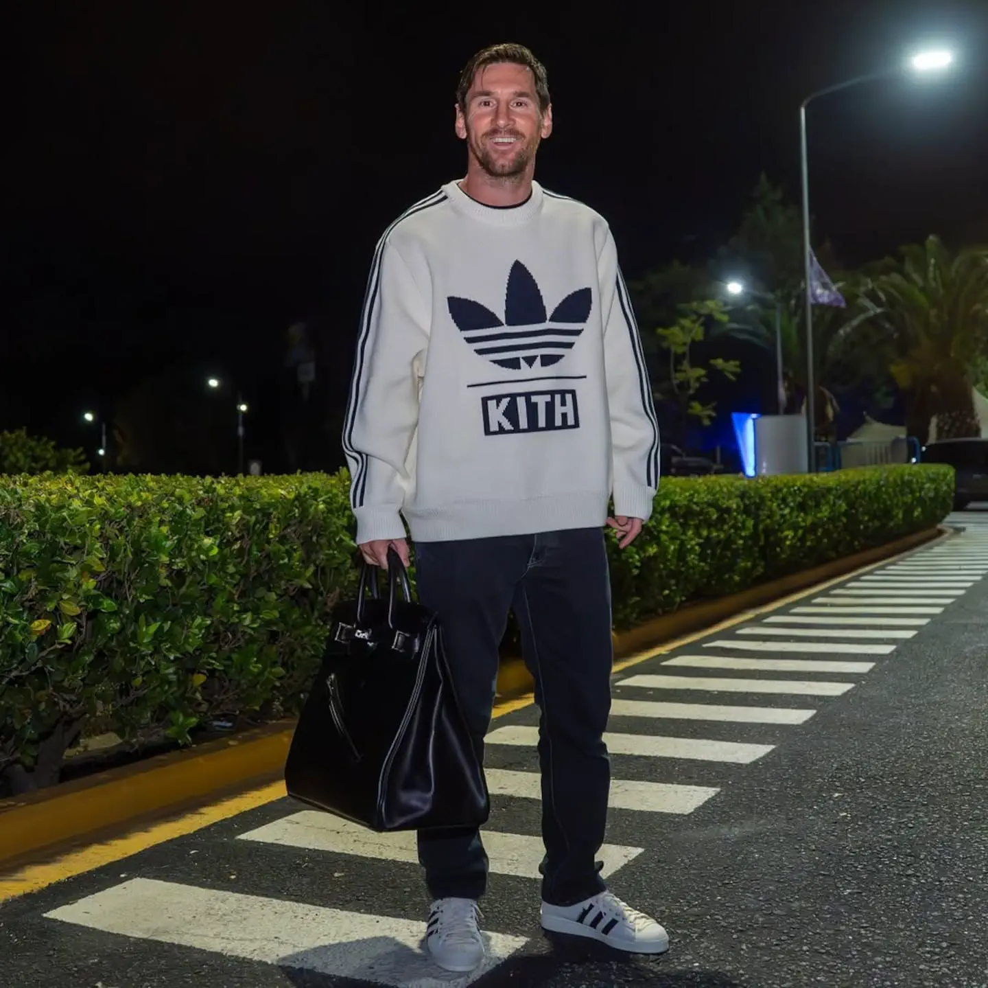 Lionel Messi parado de noche sobre una senda peatonal con un bolso de mano y buzo blanco.