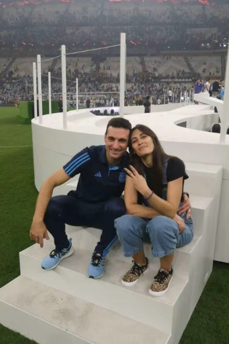 Lionel Scaloni y su esposa Elisa Montero están sentados en una tarima blanca dentro de una cancha de fútbol.