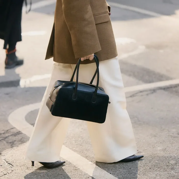 Una persona con saco marron y pantalon blanco caminando por la calle mientras lleva una cartera negra en la mano.