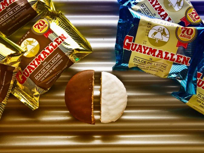 Dos mitades de alfajor, una negra y otra blanca, sobre una superficie metálica junto a paquetes de Guaymallén.