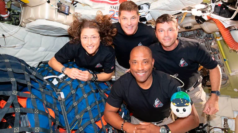 Cuatro astronautas con remera negra posan sonriendo dentro de la cabina de una nave espacial.