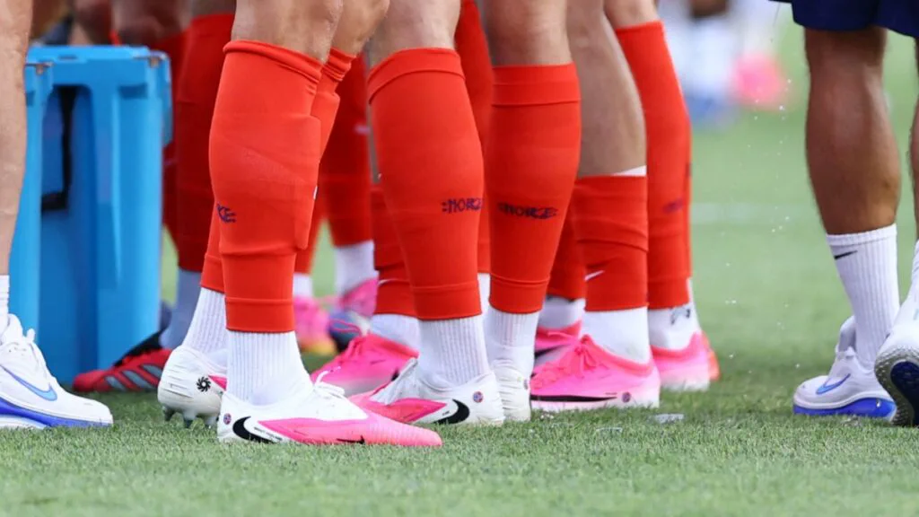 Varios futbolistas parados en el césped muestran sus medias rojas y botines rosados y blancos.