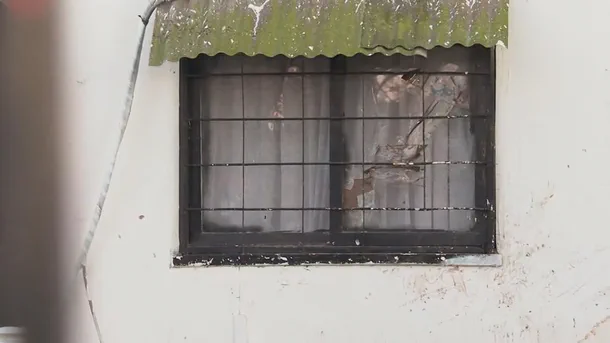 Una ventana con rejas negras y un vidrio roto se encuentra bajo un alero verde en una pared blanca.