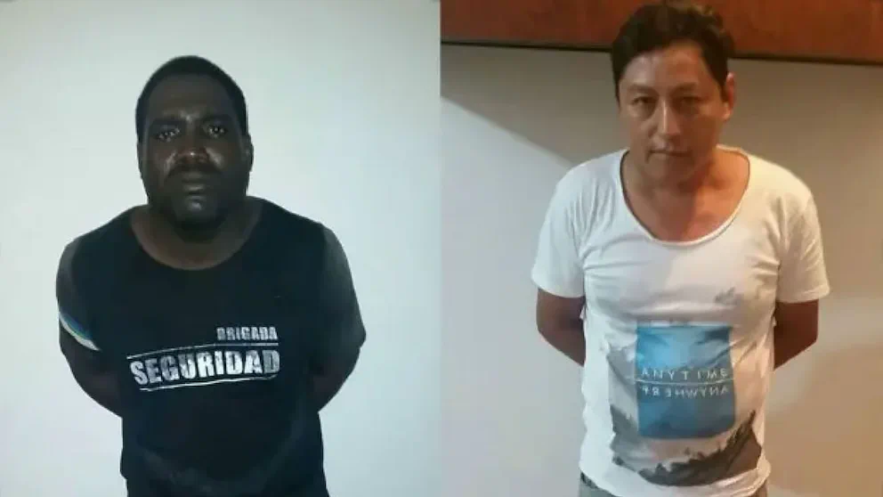 Dos hombres detenidos posan de pie con las manos detras de la espalda frente a una pared clara.