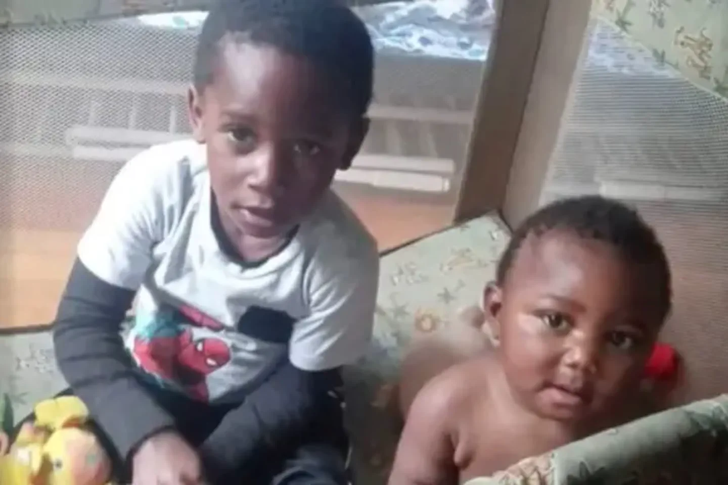 Dos nenes estan sentados dentro de un corralito de tela infantil mirando a la camara.