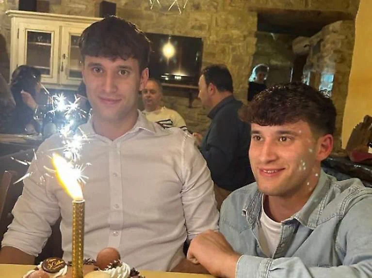 Dos muchachos sonríen sentados frente a una torta que tiene una bengala encendida.