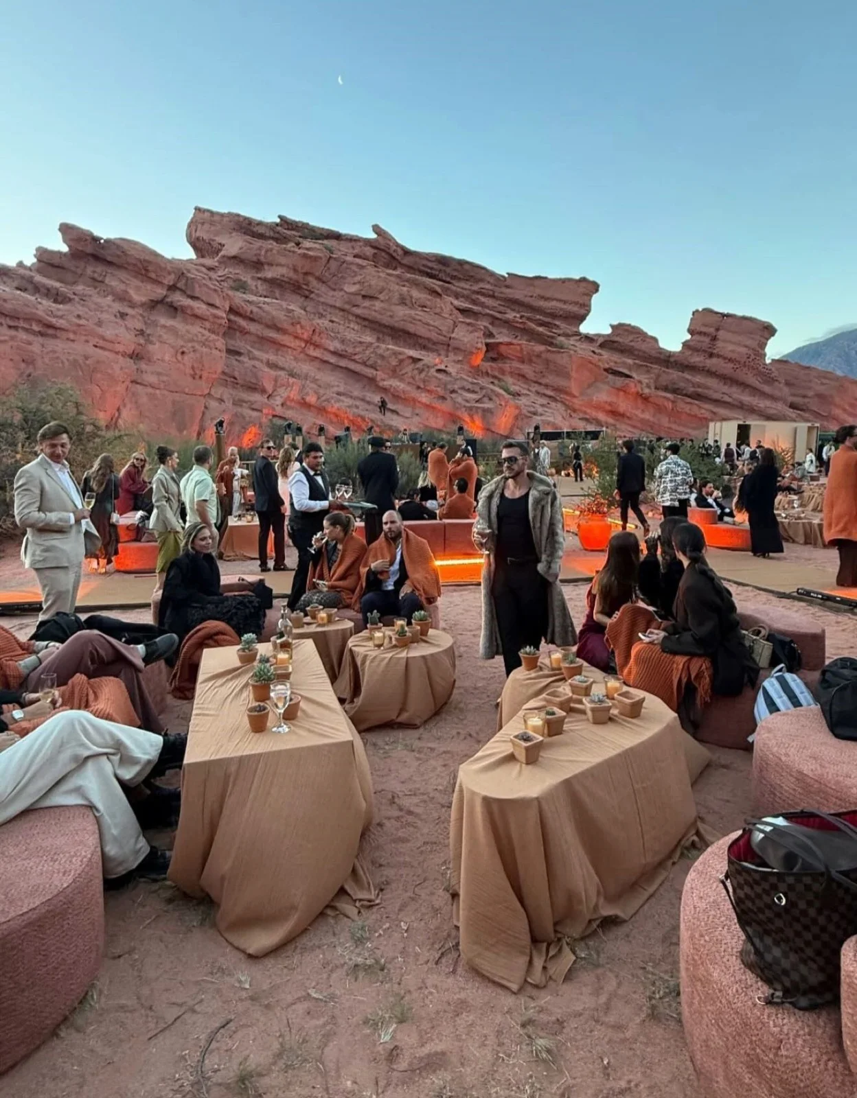 Personas reunidas en mesas bajas vestidas de gala en un paisaje desértico rodeado por formaciones rocosas rojizas.