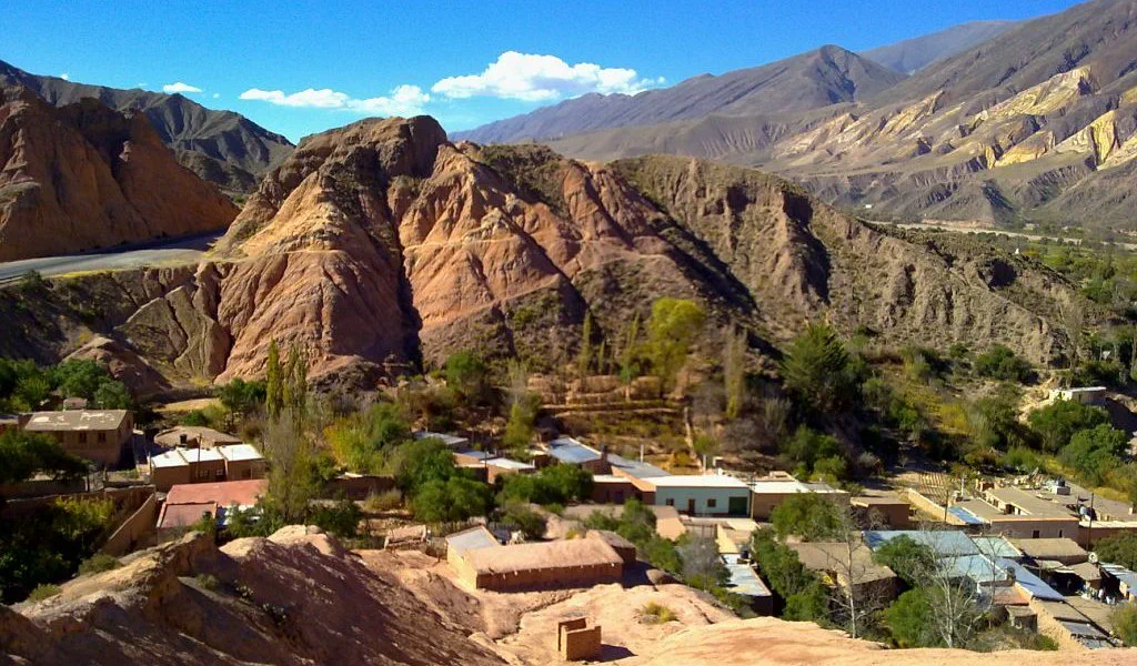Casas bajas y vegetación al pie de cerros rojizos y montañosos bajo un cielo despejado.