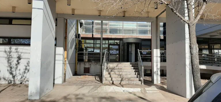 La entrada de un edificio institucional con escaleras de acceso y una puerta giratoria bajo columnas de hormigón.