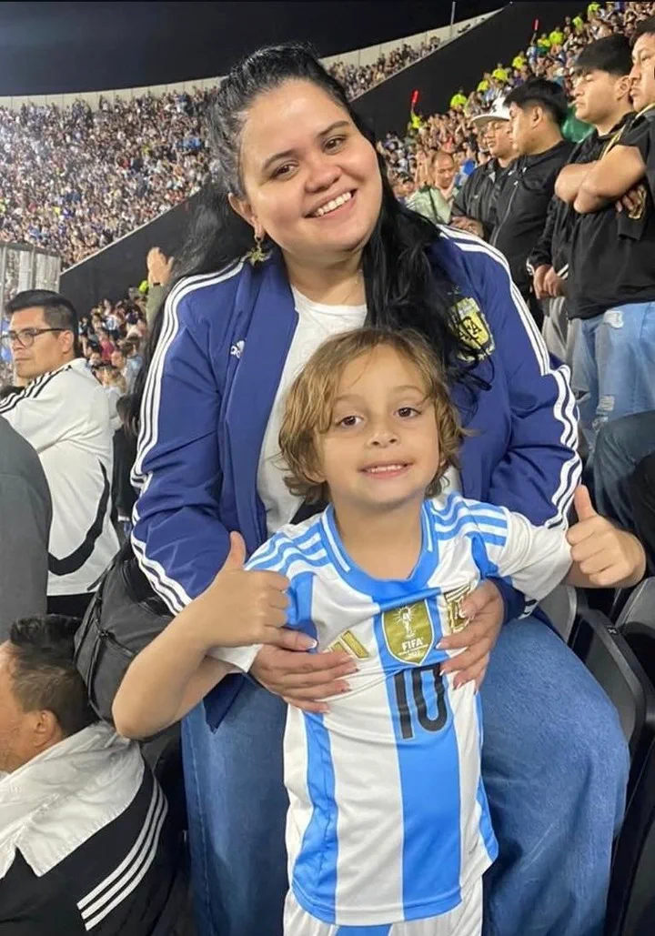 Una mujer y un nene con la camiseta de la selección argentina sonríen en la tribuna de una cancha colmada.