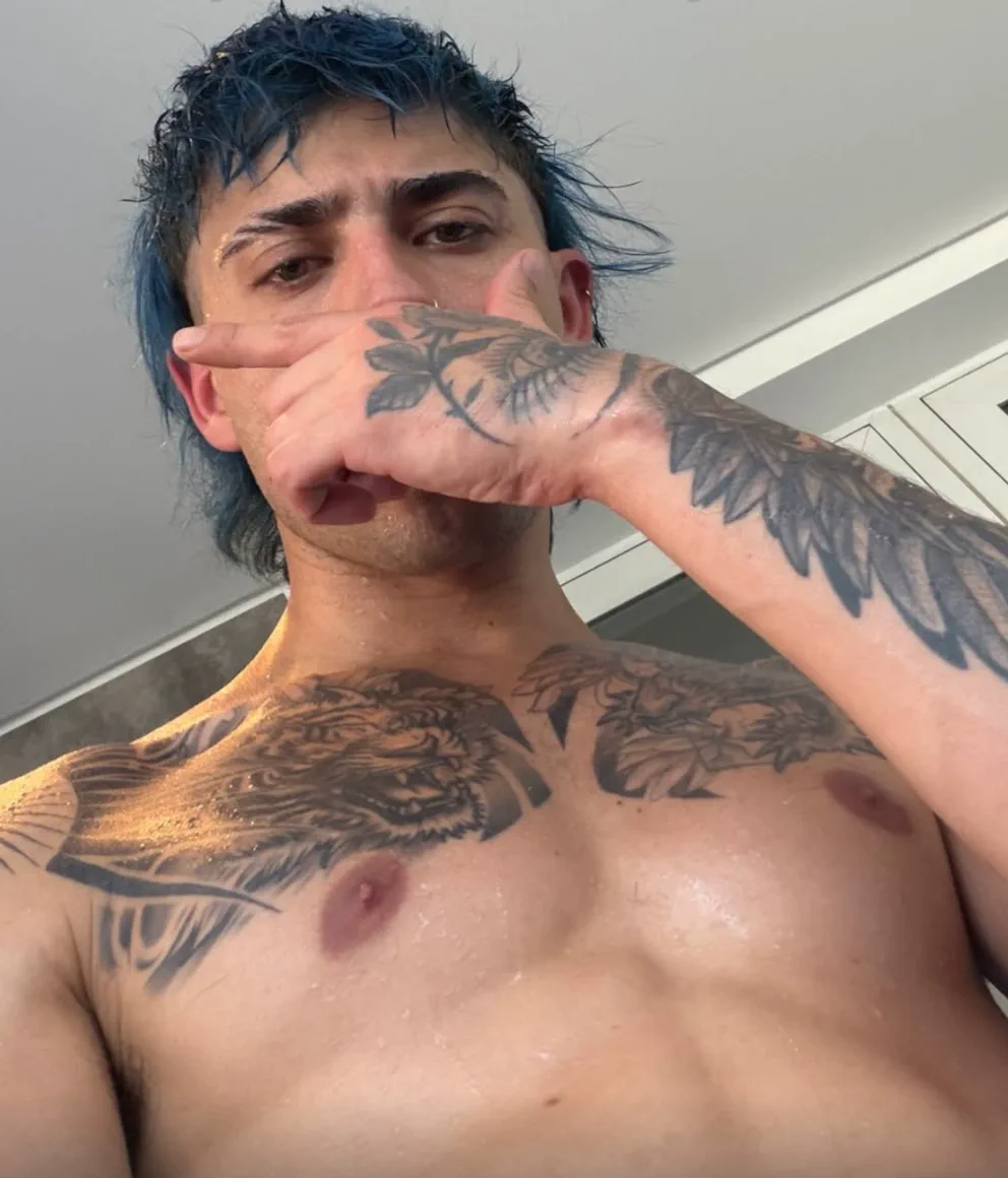 Un pibe de pelo azul y con tatuajes en el pecho y el brazo se tapa la boca con la mano.