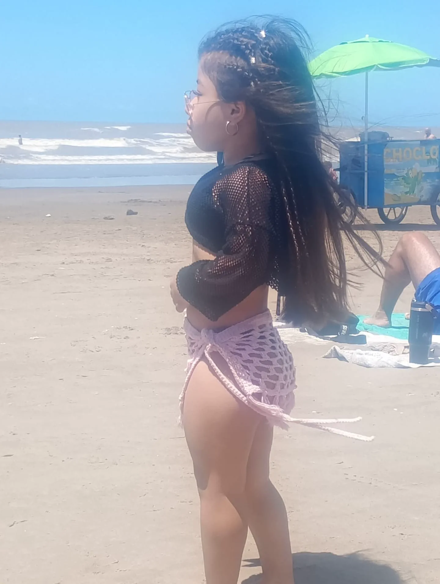 Una chica de baja estatura con ropa tejida está parada de perfil en la arena de una playa junto al mar.