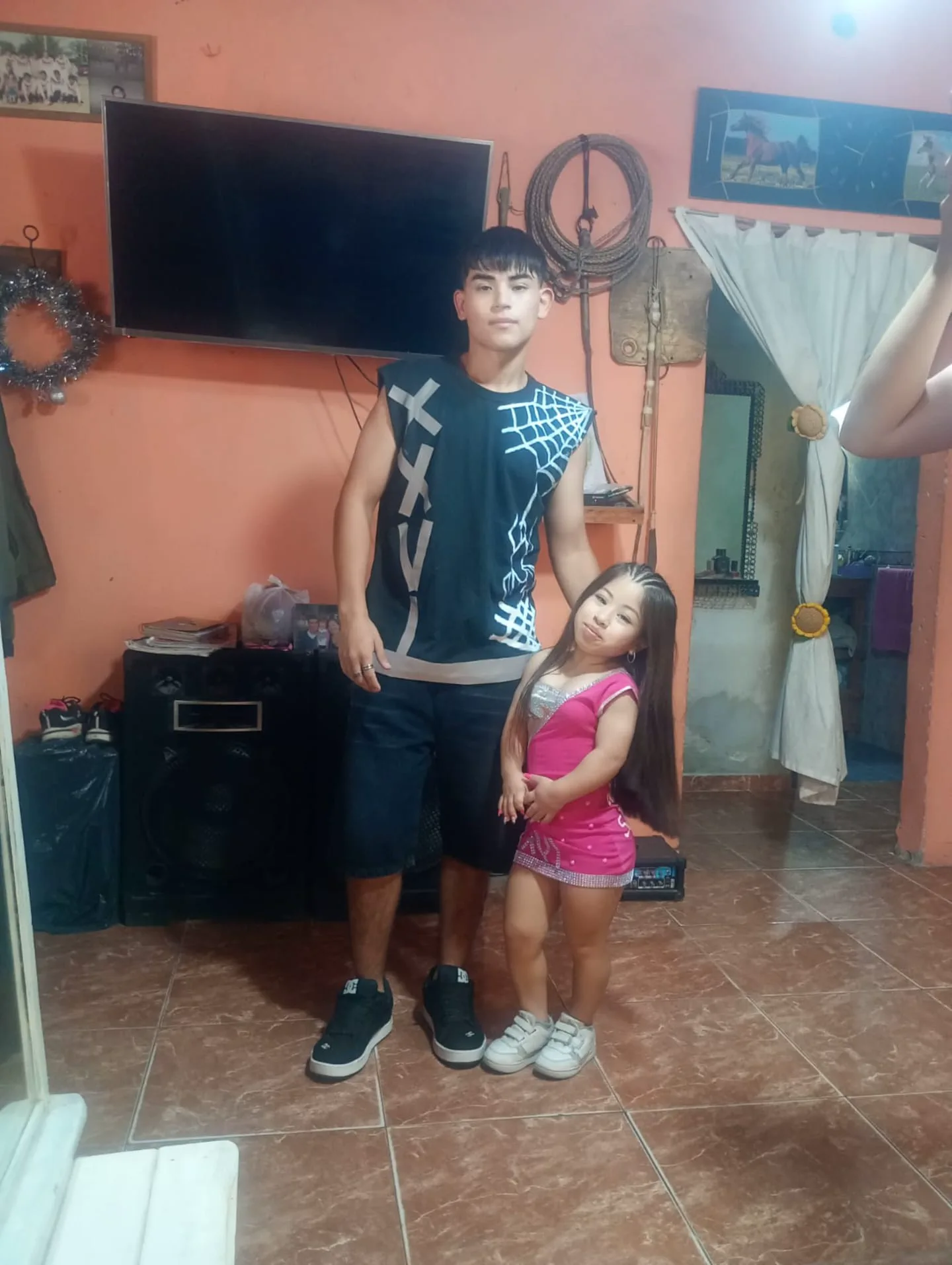 Un muchacho y una chica de baja estatura con vestido rosa están parados en el living de una casa.