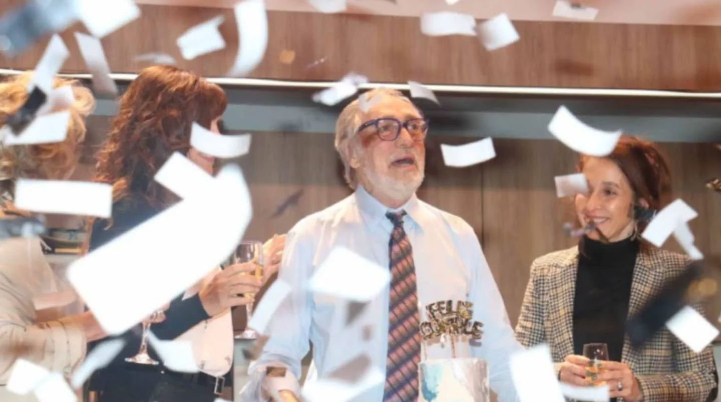 Luis Brandoni festeja su cumpleaños con una torta mientras cae papel picado alrededor junto a dos mujeres.