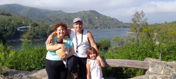 Una pareja y dos nenes posan sonrientes al aire libre con un lago y montanas de fondo.