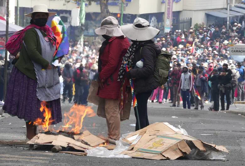 Tres personas con sombrero y barbijo están paradas en la calle frente a cartones encendidos durante una manifestación.