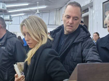 Manuel Adorni con campera oscura parado detras de una mujer rubia en un recinto cerrado.