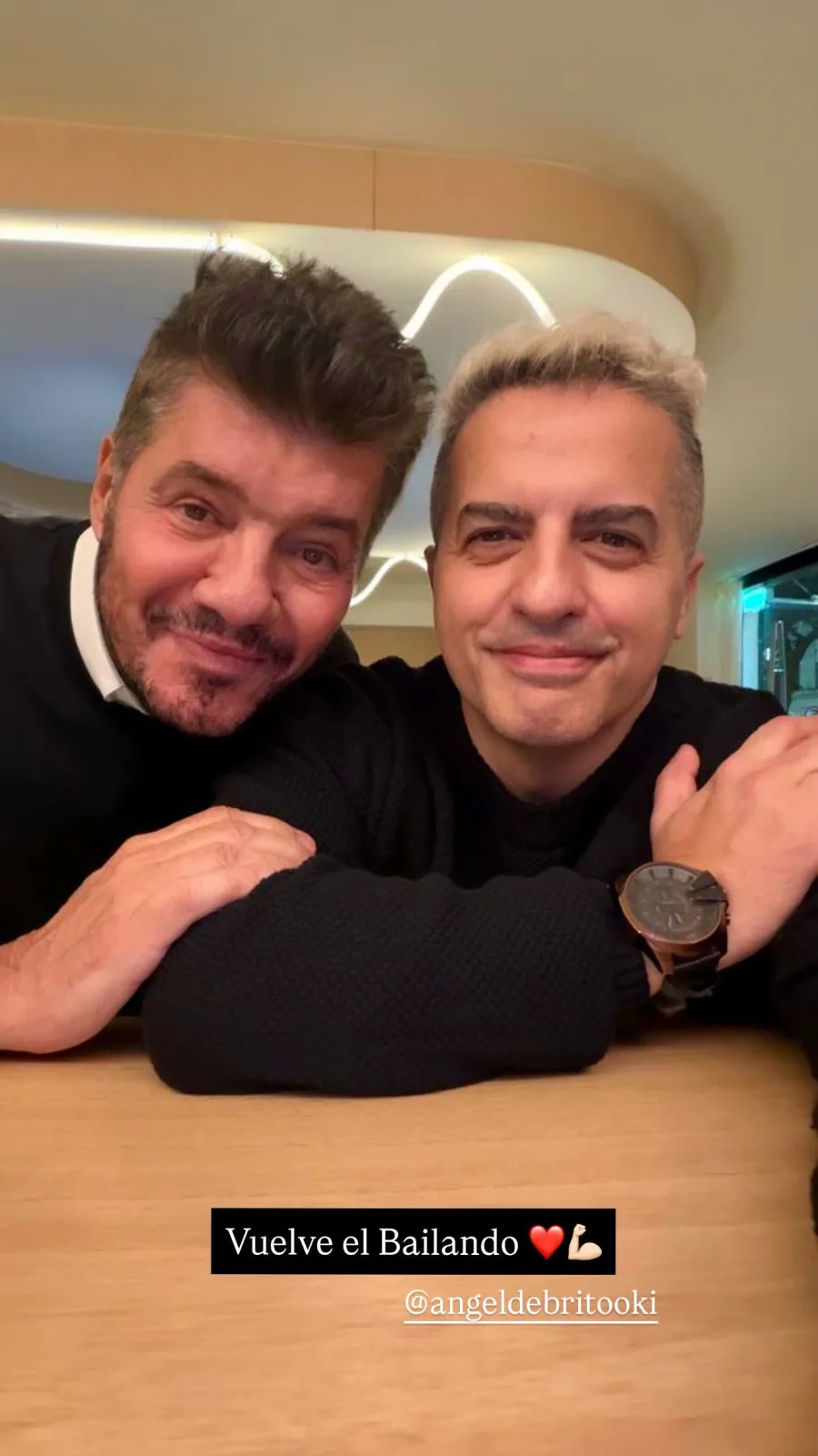 Marcelo Tinelli y Angel de Brito posan juntos sonriendo apoyados en una mesa.