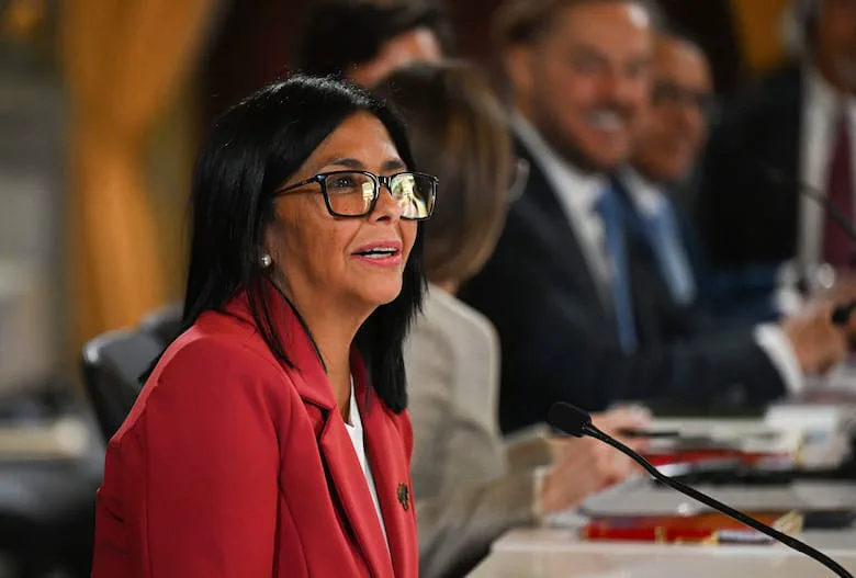 Una mujer con saco rojo y anteojos habla frente a un micrófono en una mesa de conferencia.