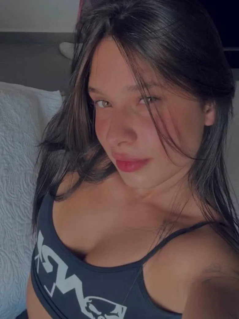 Una joven de pelo castano oscuro se saca una selfie recostada con un top deportivo negro.