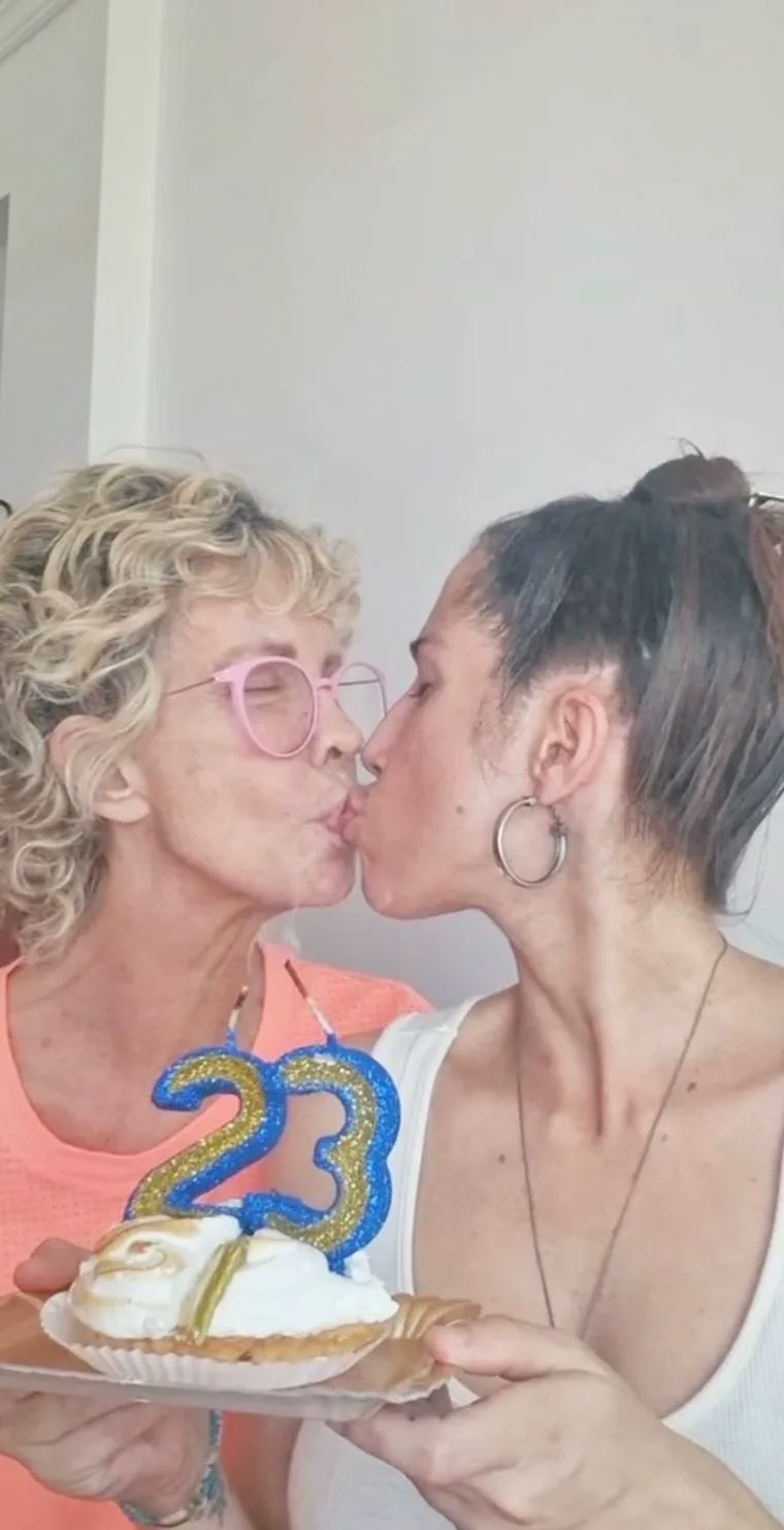 Dos mujeres se dan un beso en la boca mientras sostienen una tarta dulce con una vela con el número 23.
