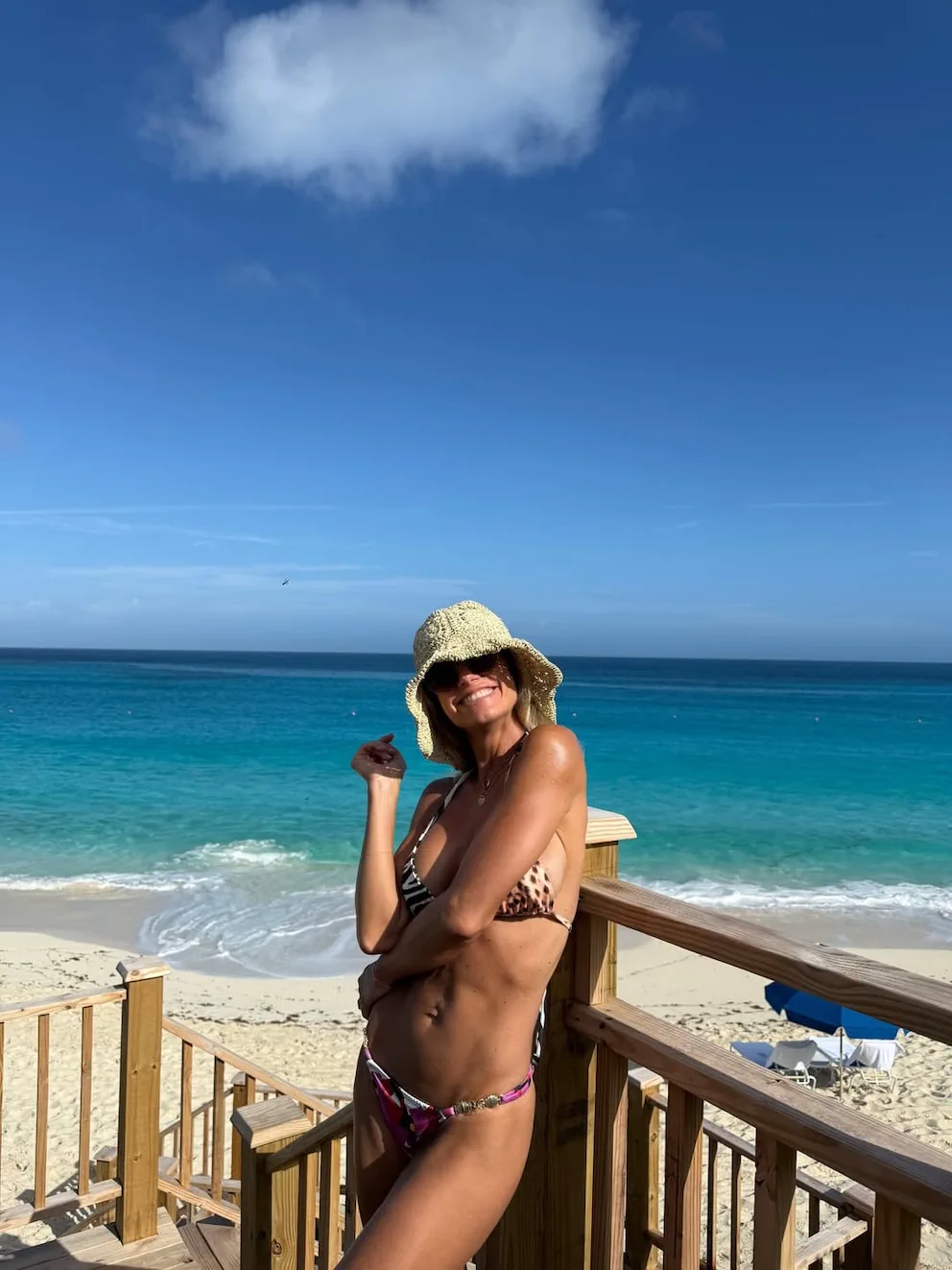 Una mujer en bikini y sombrero de paja sonríe apoyada en la baranda de madera de una escalera en la playa.