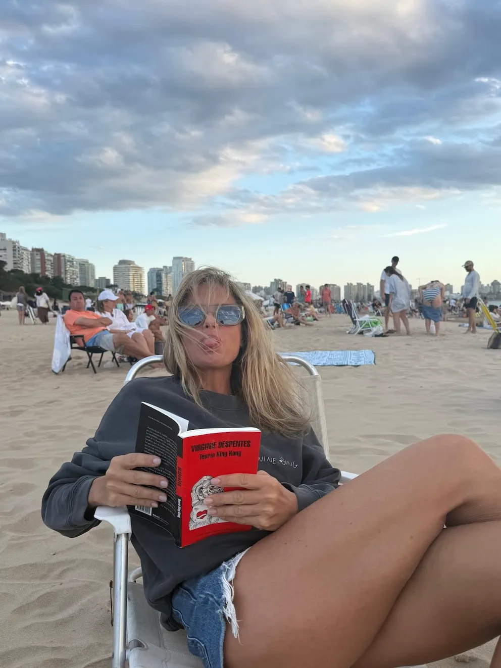 Una mujer rubia con lentes de sol saca la lengua sentada en una reposera en la playa mientras sostiene un libro.