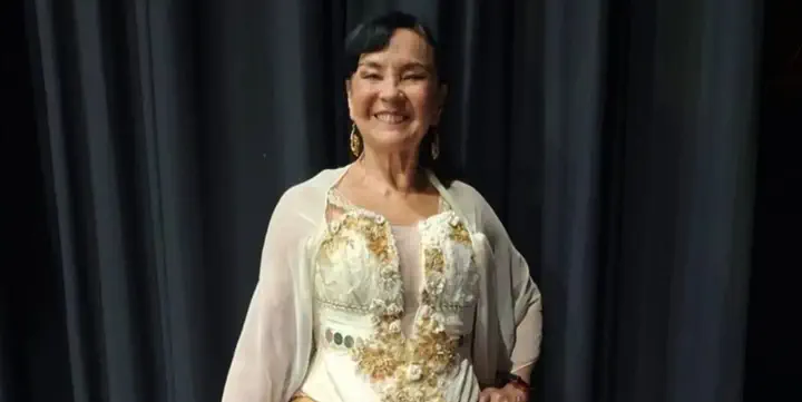 Una mujer con vestido blanco y dorado sonríe parada frente a un telón negro.