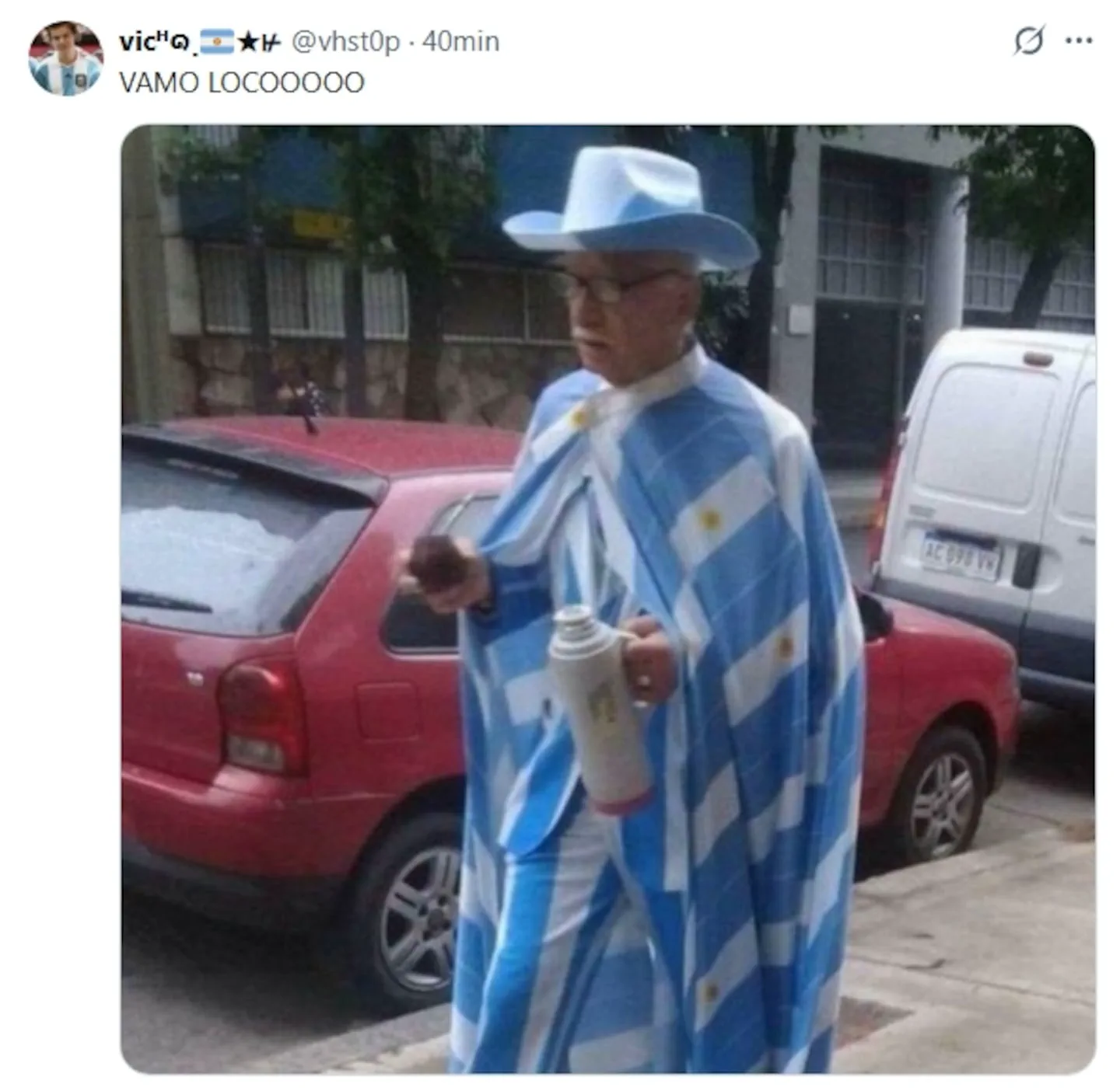 Un hombre mayor en la calle vestido con traje, poncho celeste y blanco y sombrero, llevando un mate y un termo.