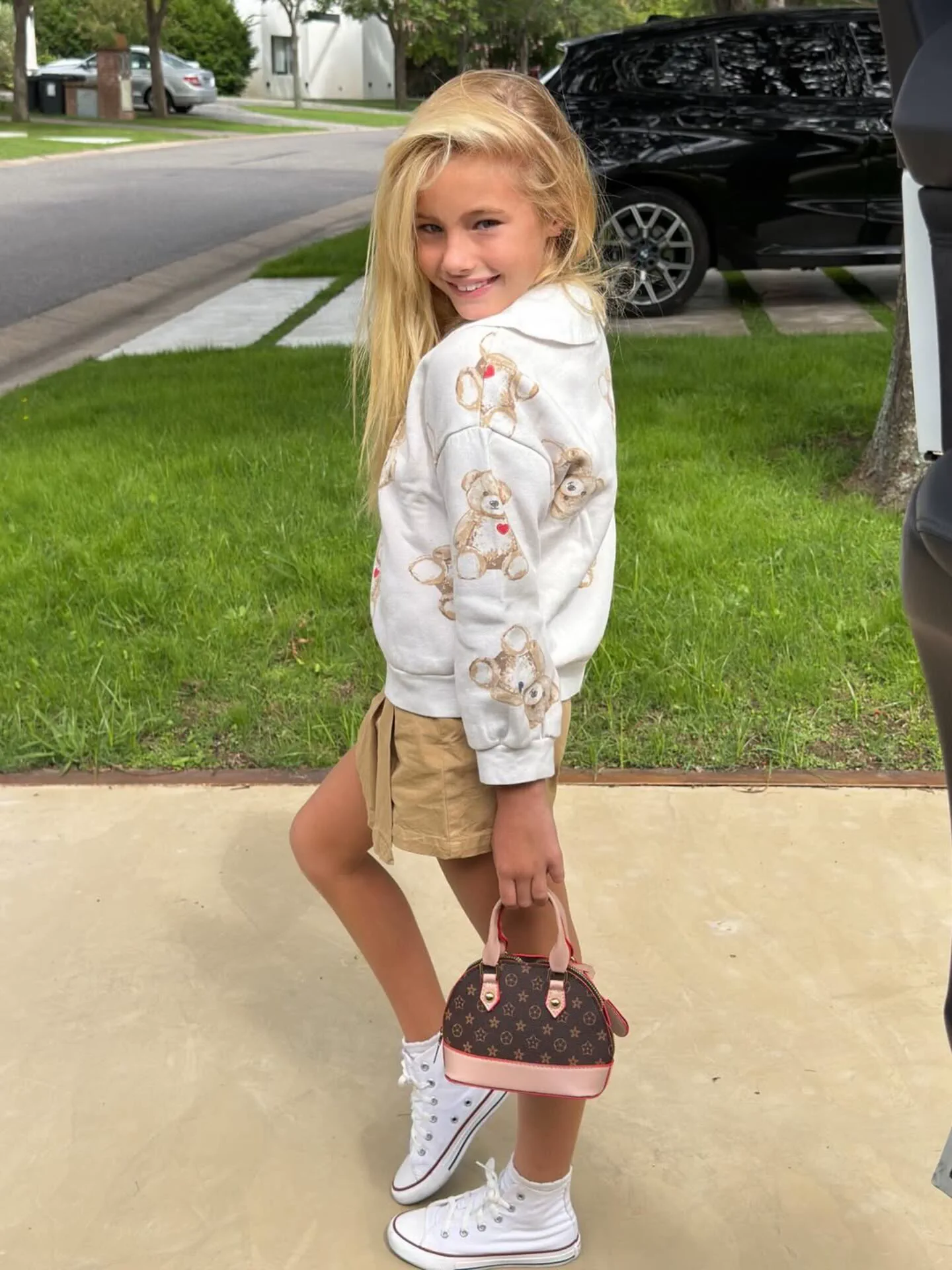 Una nena rubia con buzo blanco de ositos y pollera beige posa sosteniendo una cartera chica.