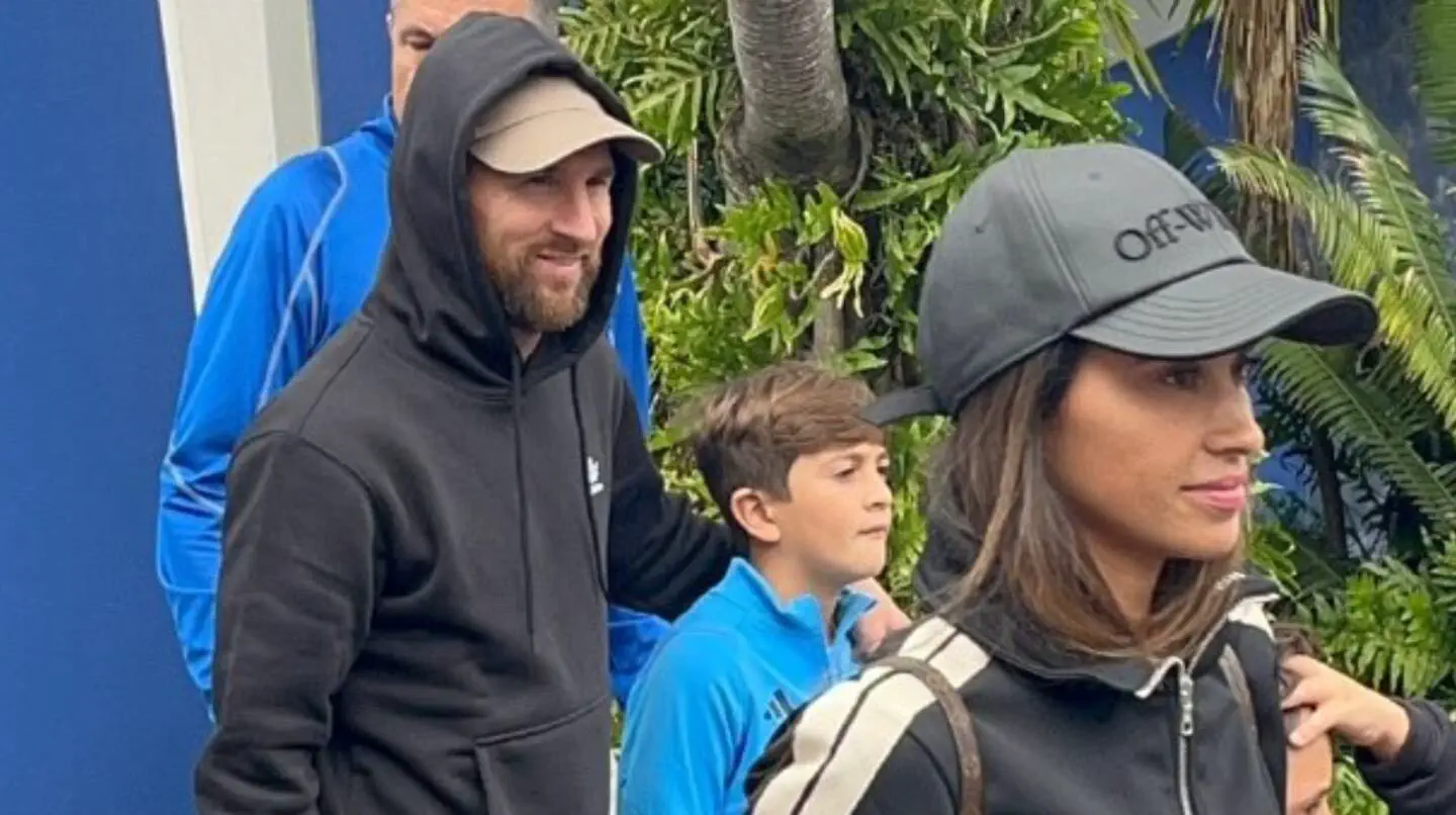 Lionel Messi camina encapuchado junto a Antonela Roccuzzo y su hijo cerca de unas palmeras.