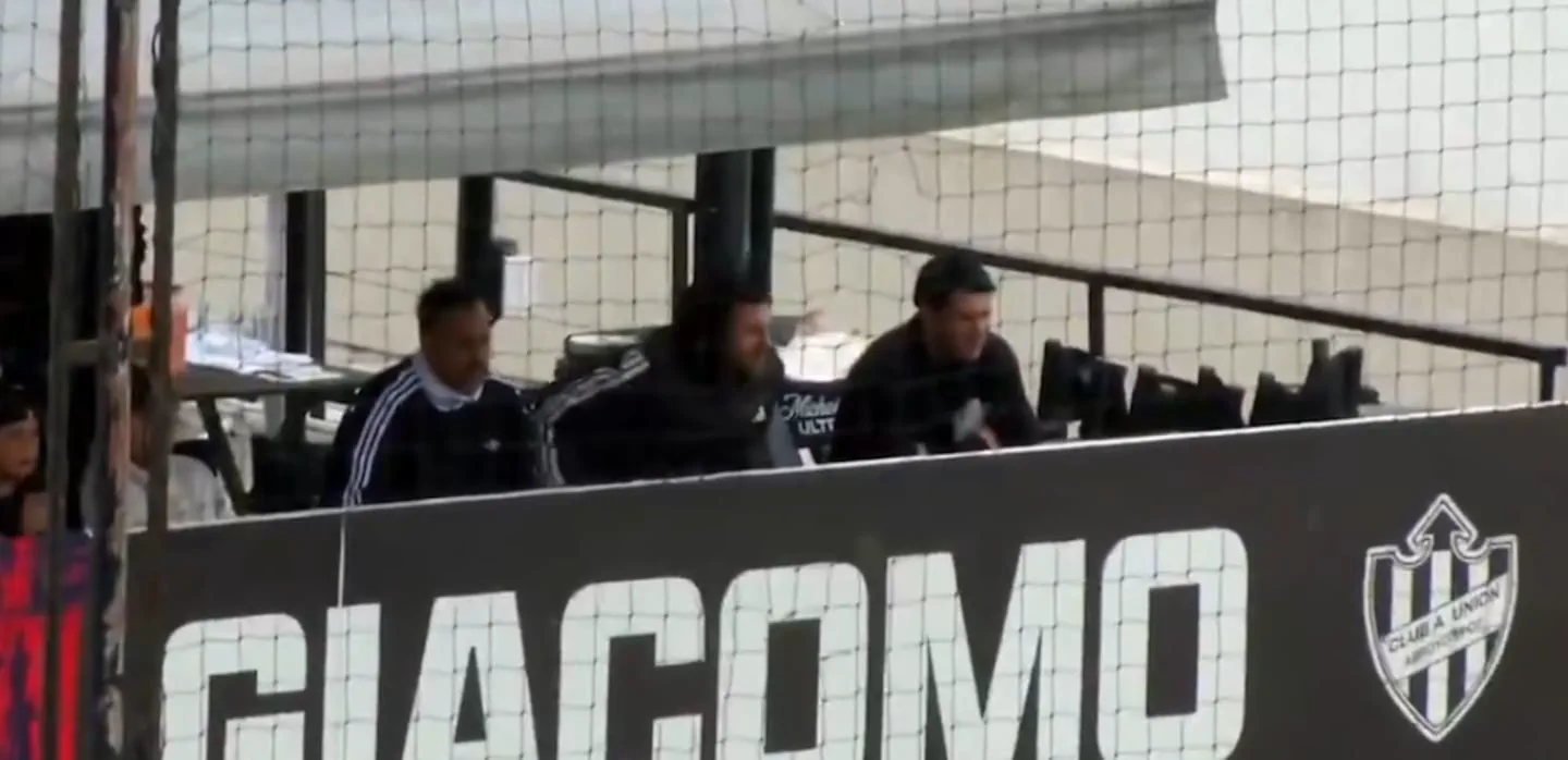 Tres hombres sentados detrás de un panel de publicidad y una red en una tribuna de una cancha deportiva.