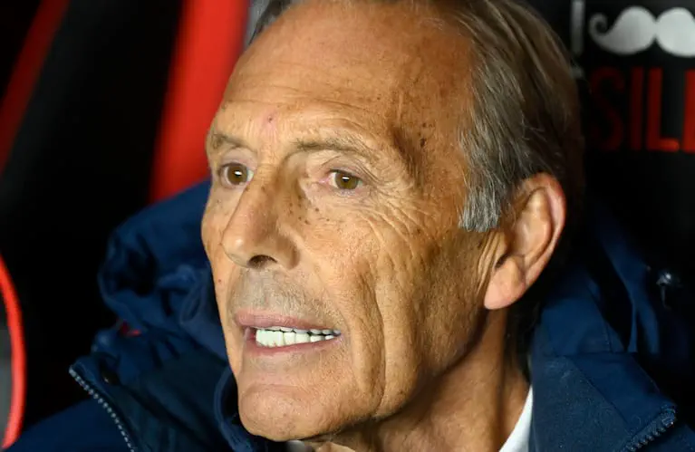 Miguel Angel Russo mira de perfil hacia un costado con campera oscura en un banco de suplentes.