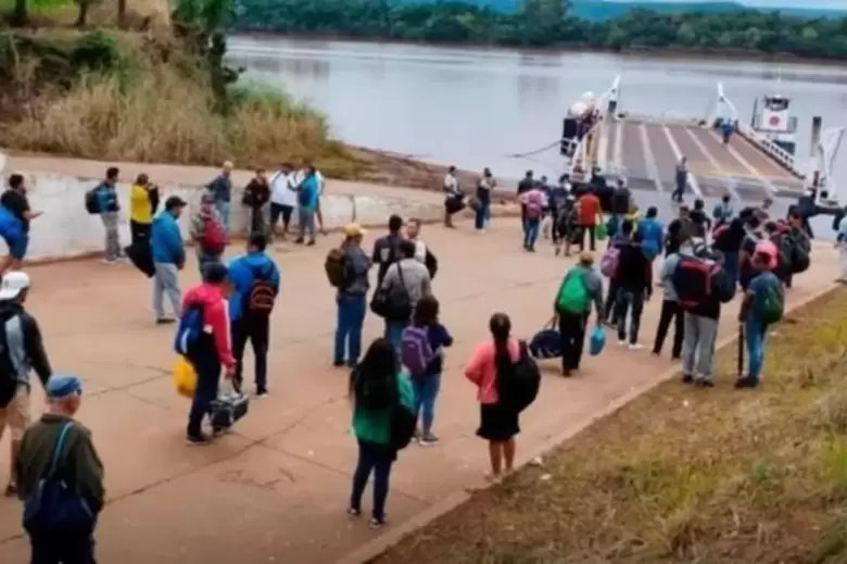 Un grupo de personas con bolsos y mochilas hace fila en una rampa de cemento para subir a una balsa en el rio.