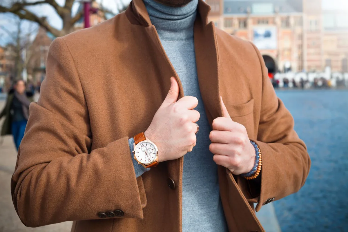Un hombre con tapado marron y polera gris muestra su reloj pulsera al aire libre.