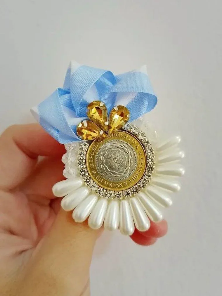 Una mano sosteniendo una escarapela artesanal con una moneda argentina, piedras brillantes y cintas celestes y blancas.