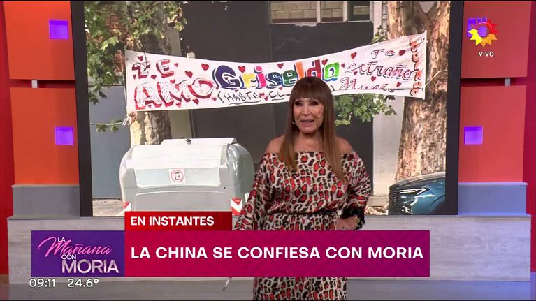 Moria Casan conduce un programa de television frente a una pantalla con un pasacalles y graficos en pantalla.